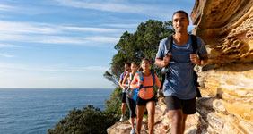 A group of four people hiking along a narrow trail on the edge of a cliff with the ocean visible in the background. The hikers are wearing backpacks and athletic clothing, walking in single file. The sky is clear with some clouds, and there are trees and vegetation along the trail.